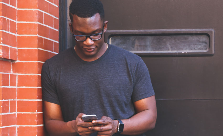 man with wearing short sleeved dark grey shirt, leaning against brick wall and looking at phone