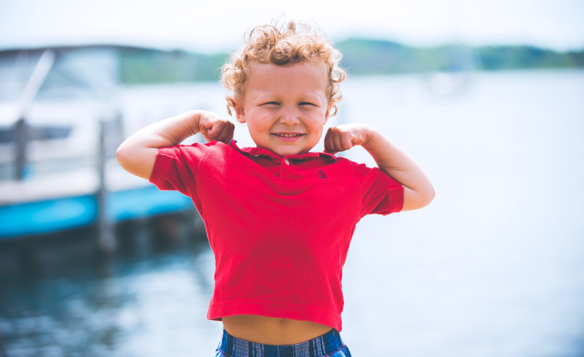 Curly-haired boy in a red polo smiles as he flexes his arms in a power pose by a lake